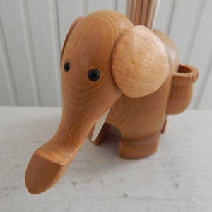 Puede incluir: Un porta palillos de dientes de madera con forma de elefante con una ranura para los palillos y un peque&ntilde;o agujero para que los palillos salgan. El elefante es marr&oacute;n y tiene ojos negros y colmillos blancos.