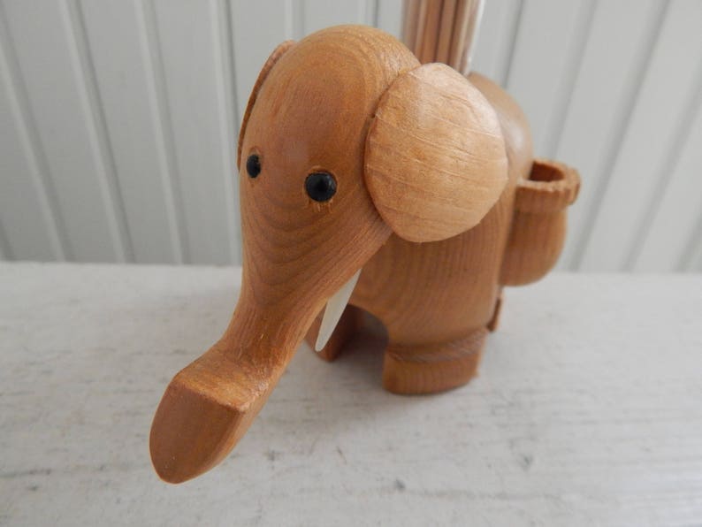 Puede incluir: Un porta palillos de dientes de madera con forma de elefante con una ranura para los palillos y un peque&ntilde;o agujero para que los palillos salgan. El elefante es marr&oacute;n y tiene ojos negros y colmillos blancos.