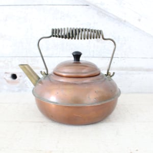 May include: A copper tea kettle with a brass spout and handle. The handle is a spring coil design. The kettle has a black knob on the lid.