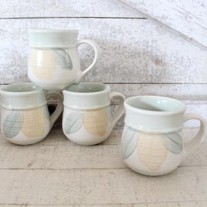 May include: Set of four ceramic mugs with a light green and yellow lemon design. The mugs have a rounded shape and a handle.
