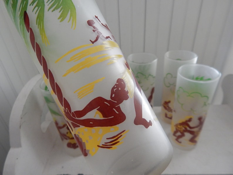 K&ouml;nnte beinhalten: Set aus mattierten Gl&auml;sern mit tropischem Insel-Motiv. Die Gl&auml;ser zeigen eine braune Silhouette einer Person auf einem Surfbrett mit einer Palme und gelben Wellen im Hintergrund.