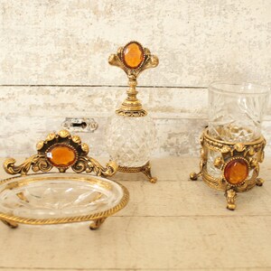 May include: A set of three vintage glass and gold bathroom accessories. The set includes a soap dish, a glass tumbler, and a bottle with a stopper. All three pieces feature a gold metal base and a clear glass body with intricate cut designs. Each piece has a large amber-colored gemstone on top.