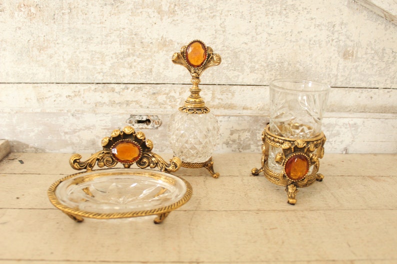 May include: A set of three vintage glass and gold bathroom accessories. The set includes a soap dish, a glass tumbler, and a bottle with a stopper. All three pieces feature a gold metal base and a clear glass body with intricate cut designs. Each piece has a large amber-colored gemstone on top.