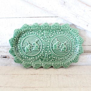 May include: A green ceramic serving dish with a scalloped edge and two raised designs of bunnies. The dish is decorated with a floral pattern.