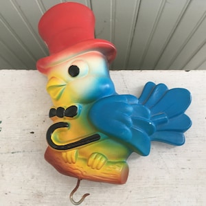 May include: A colorful plastic bird figurine with a red top hat, a black bow tie, and a blue tail. The bird is perched on a brown branch and has a metal hook on the bottom.