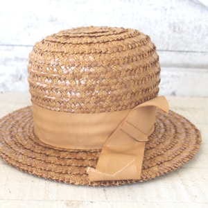 May include: A straw hat with a wide brim and a tan ribbon. The hat has a rounded crown and a woven texture. The ribbon is tied in a bow on the side. A classic accessory for sun protection and style.