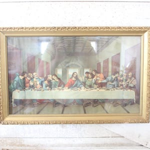 May include: A framed print of The Last Supper featuring a central figure in a red robe seated at a long table with twelve other figures. The artwork is enclosed in a rectangular gold-toned frame with a decorative, textured border pattern.