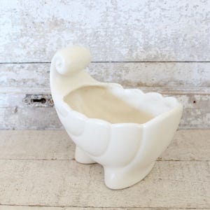 May include: A white ceramic planter shaped like a stylized shell with a rolled edge. The planter has a smooth, glossy finish and a slightly curved base.