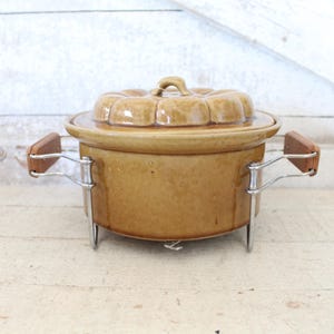 May include: A tan ceramic casserole dish with a lid, set on a metal stand with wooden handles. The lid has a decorative, scalloped design. The dish is designed for cooking and serving.