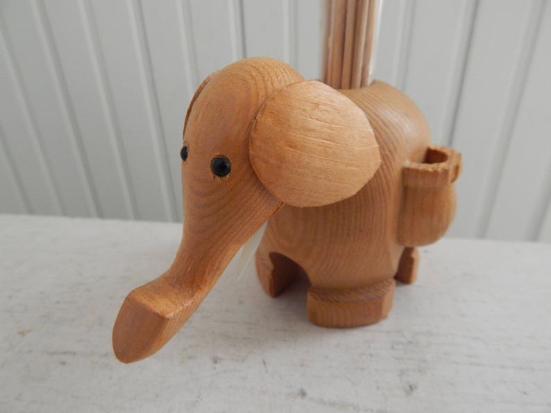 Puede incluir: Porta palillos de dientes de madera en forma de elefante con ojos negros. El elefante tiene un peque&ntilde;o compartimento en el lateral para los palillos de dientes.
