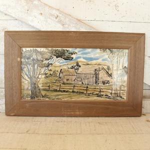 May include: A framed drawing of a farmhouse with a brown wooden frame. The drawing is in black and white and depicts a farmhouse with a barn and trees in the background.