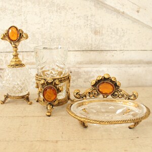 May include: A set of three vintage bathroom accessories with gold accents and amber glass accents. The set includes a bottle, a tumbler, and a soap dish.