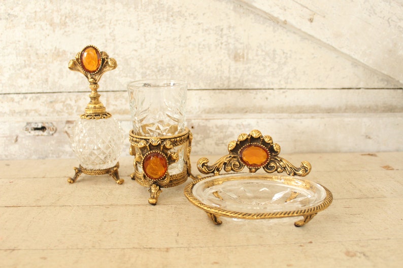 May include: A set of three vintage bathroom accessories with gold accents and amber glass accents. The set includes a bottle, a tumbler, and a soap dish.