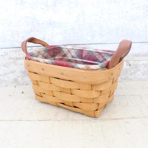 May include: A rectangular woven basket with leather handles and a plaid fabric liner. The basket is made of natural wicker and has a light brown color.