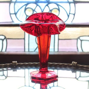May include: A vibrant red glass vase with a flared top and a hexagonal base. The vase has a long, slender stem and is set against a backdrop of a stained-glass window. The glass reflects light, creating a striking visual.