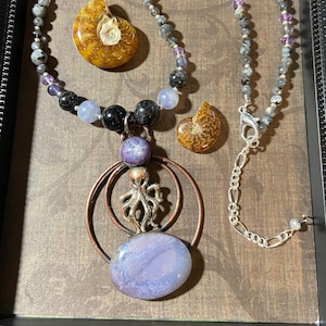 May include: A necklace with a large purple agate pendant, a silver octopus charm, and two ammonite fossils. The necklace is made with black and gray beads and purple accents.