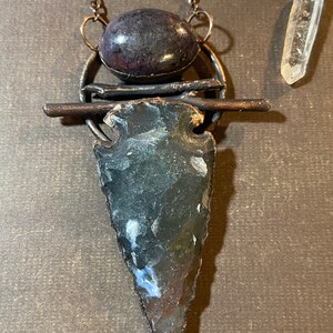 May include: A necklace with a copper chain and a pendant featuring a dark grey arrowhead, a dark purple oval stone, and a copper bar with two copper rings.