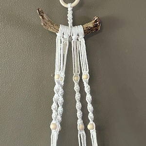 May include: White macrame plant hanger with wooden beads and a natural wood branch. The hanger is suspended from a metal hook and a wooden ring. The design features intricate knotting and a bohemian aesthetic, perfect for displaying plants.