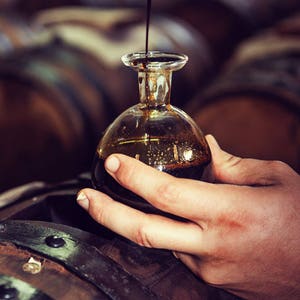 Puede incluir: Una peque&ntilde;a botella de vidrio transparente con cuerpo redondeado y cuello estrecho se llena con un l&iacute;quido oscuro. La botella se sostiene sobre una barrica de madera, con otras barricas visibles en el fondo. El l&iacute;quido parece ser vinagre bals&aacute;mico.