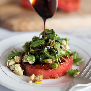 Puede incluir: Una ensalada gourmet con una gruesa rodaja de sand&iacute;a roja cubierta con r&uacute;cula, pistachos y un chorrito de glaseado bals&aacute;mico oscuro. La ensalada se sirve en un plato blanco con un tenedor plateado.