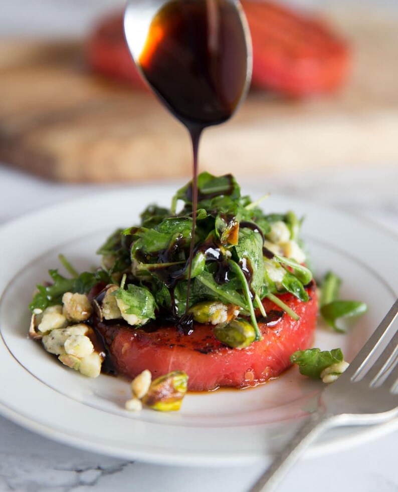 Puede incluir: Una ensalada gourmet con una gruesa rodaja de sand&iacute;a roja cubierta con r&uacute;cula, pistachos y un chorrito de glaseado bals&aacute;mico oscuro. La ensalada se sirve en un plato blanco con un tenedor plateado.