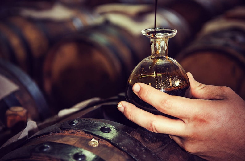 Puede incluir: Una peque&ntilde;a botella de vidrio transparente con cuerpo redondeado y cuello estrecho se llena con un l&iacute;quido oscuro. La botella se sostiene sobre una barrica de madera, con otras barricas visibles en el fondo. El l&iacute;quido parece ser vinagre bals&aacute;mico.