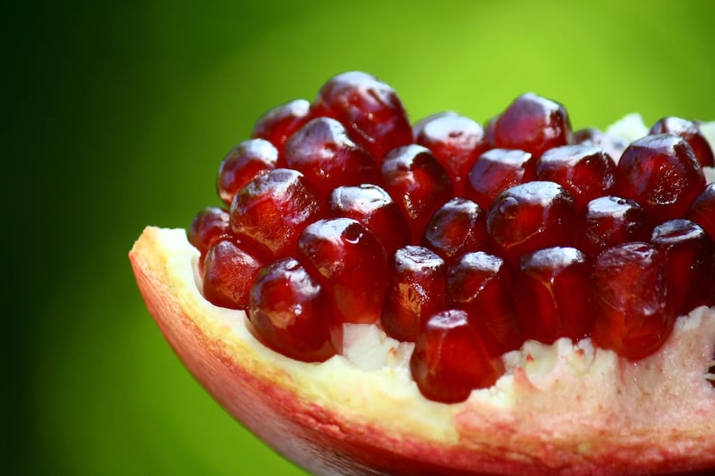 Puede incluir: Primer plano de un segmento de fruta de granada. El interior de la fruta revela numerosas semillas de color rojo intenso y brillante. La c&aacute;scara es una mezcla de rojo y blanco, con un fondo verde. La imagen destaca la textura y el color de la fruta.