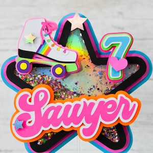 May include: A pink and orange star-shaped cake topper with a rainbow roller skate, a pink heart, and the name "Sawyer" in pink letters. The number "7" is also included in blue and green.