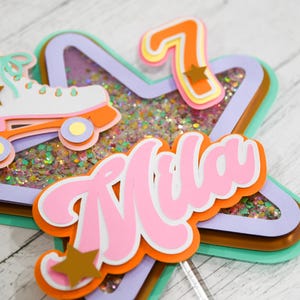 May include: A multi-layered star-shaped cake topper with a pink and orange "Mila" name, a glittery center, and a white roller skate with gold stars. The number "7" is also included in the design.