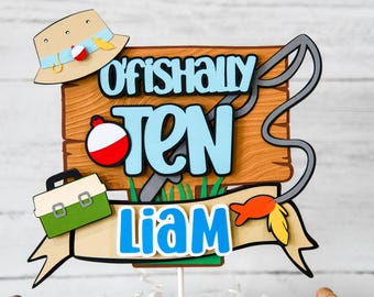 Puede incluir: Un adorno para pastel azul y blanco con el texto "Officially Ten" y una caña de pescar. El adorno también incluye una caja de aparejos verde, un flotador de pesca rojo y blanco y un pez amarillo y naranja. El nombre "Liam" está escrito en letras azules en una pancarta.