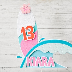May include: A colorful surfboard-shaped cake topper with the number "13" in orange and a pink hibiscus flower. The name "Kiara" is displayed in pink letters on a blue wave design. The topper is made of layered paper.