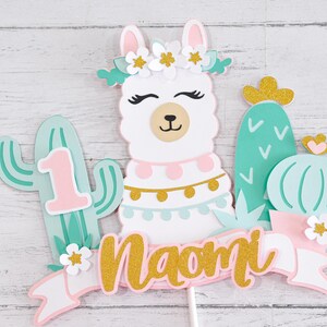 May include: A white llama cake topper with a pink and blue flower crown, gold glitter accents, and the name "Naomi" on a pink and white banner. The llama is surrounded by green cactus cutouts with white accents.