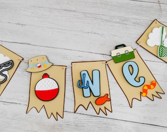 May include: A brown and tan banner with the word "ONE" in blue and white letters. The banner is decorated with fishing-themed cutouts, including a fishing rod, a fishing lure, a fishing hat, a tackle box, and a fish.
