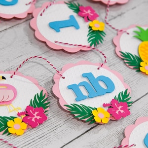 May include: Pink and white scalloped paper circles with blue letters and tropical decorations, including a flamingo, pineapple, and flowers. The circles are strung together with twine.