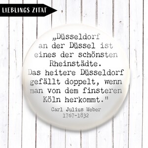 Könnte beinhalten: Ein weißer, runder Button mit dem Text "LIEBLINGS ZITAT" in einem schwarzen Pfeil. Der Button zeigt ein Zitat auf Deutsch über Düsseldorf, zugeschrieben Carl Julius Weber (1767-1832), vor einem weißen Holz Hintergrund.