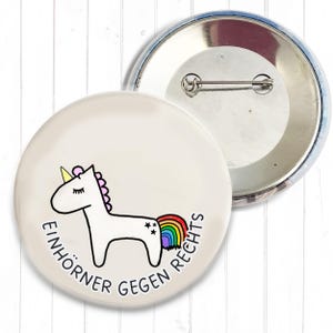 Könnte beinhalten: Ein weißer Button mit einem Einhorn-Cartoon-Design, einer Regenbogen-Schwanz, einer rosa Mähne und einem gelben Horn. Der Text "EINHÖRNER GEGEN RECHTS" ist um das Einhorn herum gedruckt. Ein zweiter Button ist im Hintergrund sichtbar.