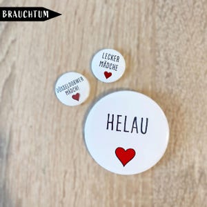 Könnte beinhalten: Drei runde weiße Ansteckbuttons in verschiedenen Größen. Der größte Button zeigt den schwarzen Schriftzug HELAU und ein rotes Herz. Die zwei kleineren Buttons zeigen den schwarzen Schriftzug LECKER MÄDCHE und DÜSSELDORWER MÄDCHE, jeweils mit einem kleinen roten Herz.