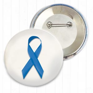 Könnte beinhalten: Ein runder Ansteckbutton mit weißem Hintergrund und einem mittig platzierten blauen Awareness-Band. Die Rückseite des Buttons besteht aus silberfarbenem Metall und verfügt über eine horizontale Nadel zum Befestigen an Stoffen oder Materialien.