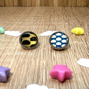 Könnte beinhalten: Zwei runde, silberumrandete Glascabochons mit unterschiedlichen Designs. Ein Cabochon hat ein schwarzes und goldenes Tiermuster. Der andere Cabochon hat einen blauen Hintergrund mit weißen Wolken.
