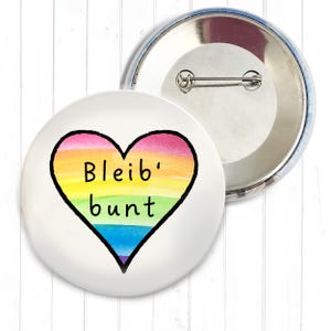 Könnte beinhalten: Ein weißer Ansteckbutton mit einem Regenbogenherz-Design und den schwarzen Worten "Bleib' bunt". Das Herz ist mit Regenbogenfarben gefüllt. Ein zweiter Button ist im Hintergrund zu sehen.
