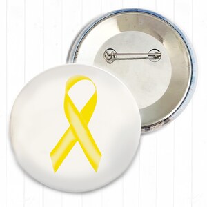 Könnte beinhalten: Zwei runde Ansteckbuttons. Einer zeigt ein gelbes Band auf weißem Hintergrund. Der andere ist silberfarben mit einem Nadelverschluss. Das gelbe Band ist ein Symbol für Unterstützung und Bewusstsein.