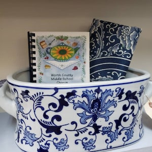 May include: A white ceramic planter with blue floral designs and two handles. The planter is filled with a blue and white patterned fabric and a small spiral bound book with the text "Worth County Middle School Chorus".