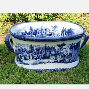 May include: A large, oval-shaped ceramic planter with a blue and white transferware design. The planter features a detailed landscape scene with buildings, trees, and figures. It has two blue handles and sits on a grassy surface.