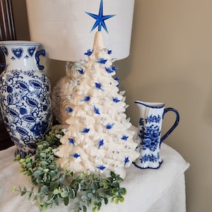May include: A white ceramic Christmas tree with blue bird ornaments and a blue star on top. The tree is decorated with white lights and is sitting on a white table with a blue and white vase and pitcher.