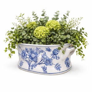 May include: Oval white ceramic planter with blue floral design and handles, filled with green foliage and chartreuse flowers. The planter is a decorative piece for indoor or outdoor use.