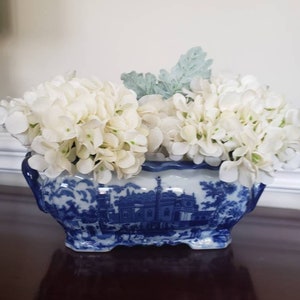 May include: A blue and white ceramic planter with a landscape design, filled with white faux hydrangea flowers and a few green leaves. The planter has a scalloped rim and is sitting on a dark brown surface.