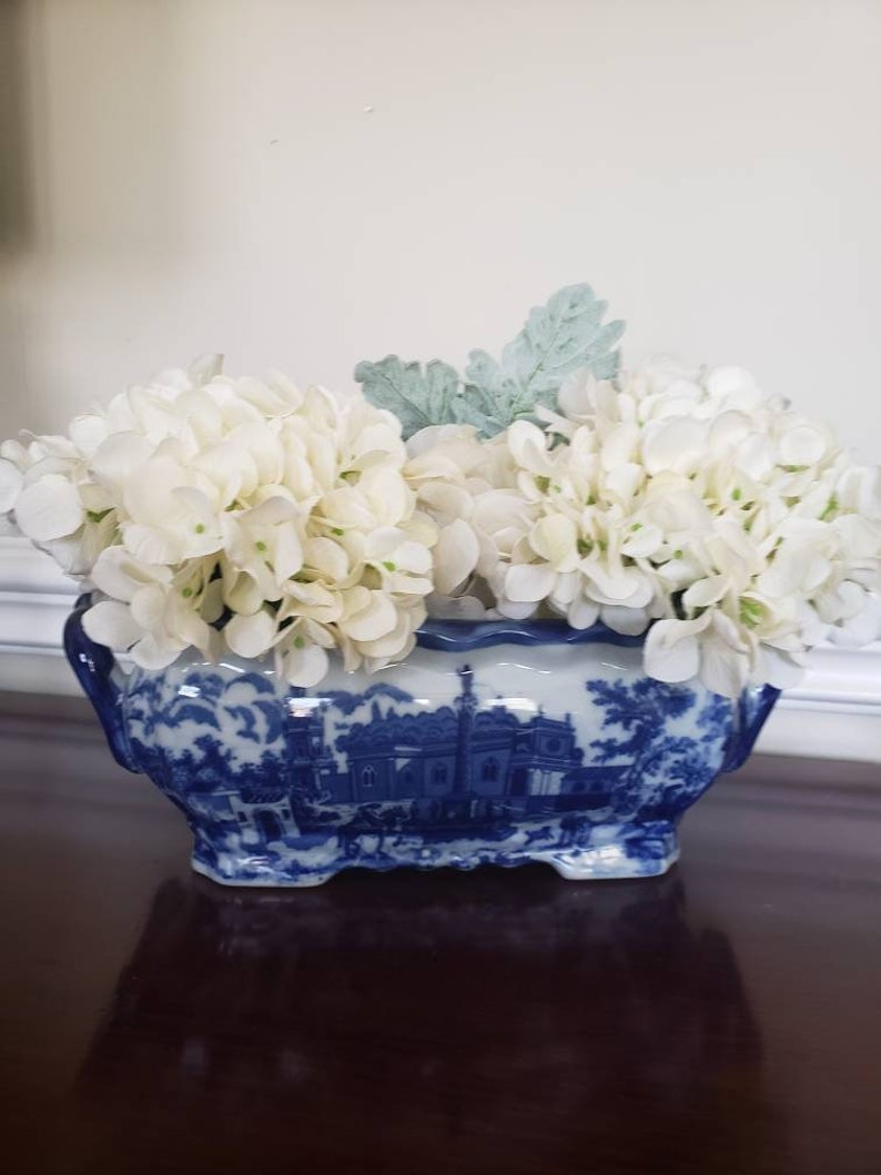 May include: A blue and white ceramic planter with a landscape design, filled with white faux hydrangea flowers and a few green leaves. The planter has a scalloped rim and is sitting on a dark brown surface.