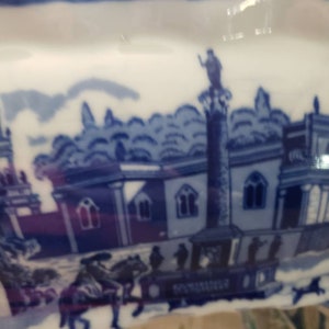 May include: A white and blue ceramic dish with a scalloped edge. The dish features a blue and white transferware design depicting a historical scene with buildings, figures, and a horse-drawn carriage. The rim is a solid blue.