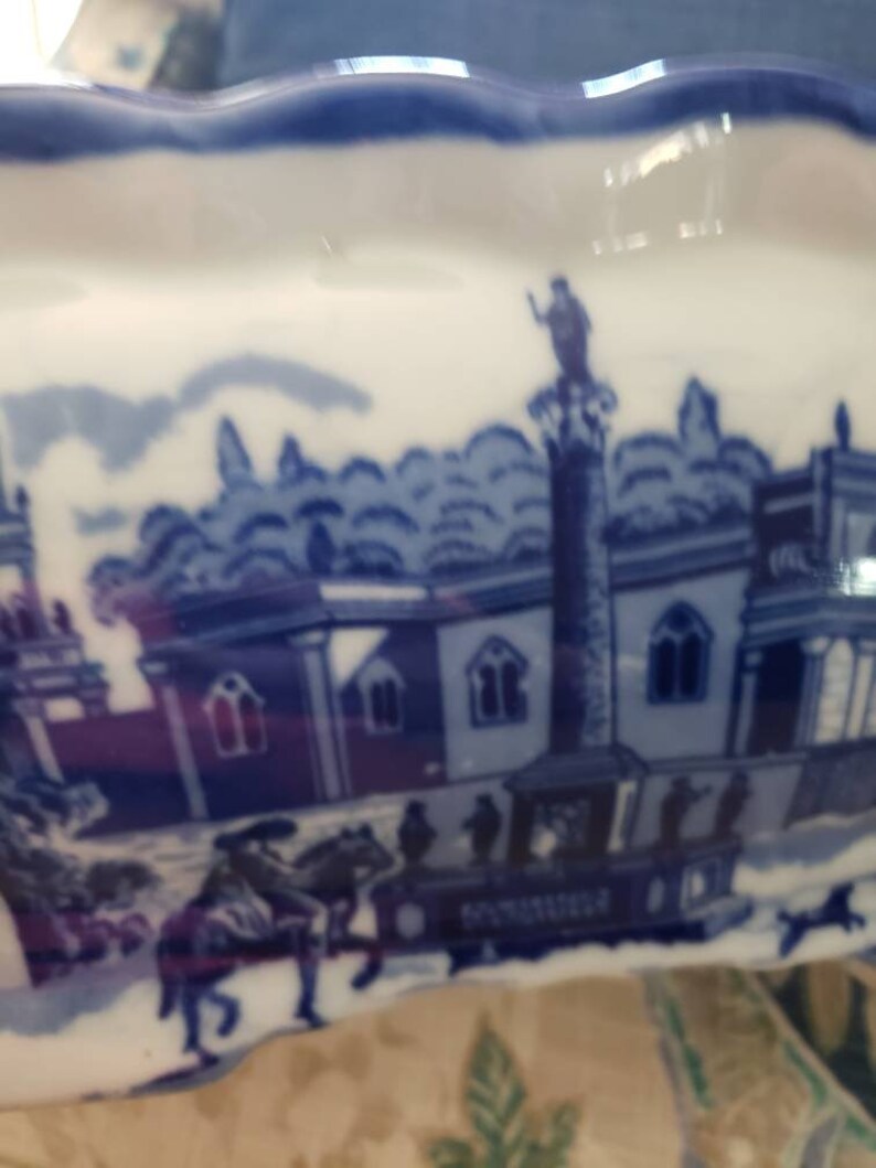 May include: A white and blue ceramic dish with a scalloped edge. The dish features a blue and white transferware design depicting a historical scene with buildings, figures, and a horse-drawn carriage. The rim is a solid blue.