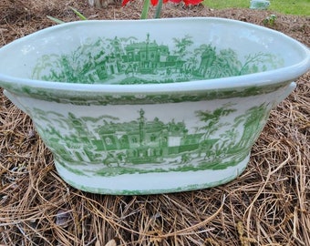 Könnte beinhalten: Ovale, cremefarbene Keramikschale mit grünem Transferdruck-Landschaftsmotiv. Die Schale steht auf Kiefernnadeln, mit roten Blumen im Hintergrund. Die Schale hat einen breiten Rand und eine leicht geschwungene Form. Das Design ist im Toile-de-Jouy-Stil.